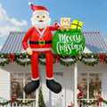 Santa inflatable sitting on a porch roof with a green Merry Christmas sack and a teddy bear