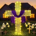 Lighted cross draped with a purple sash standing among white lilies at dusk