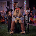 Sitting scarecrow prop with red eyes holding a candy bowl while kids in costume reach for treats